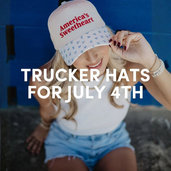 Blonde woman wearing a cream trucker hat that says “America’s Sweetheart” in red embroidery with a blue printed bill, styled with denim shorts and white tank, promoting trucker hats for July 4th.