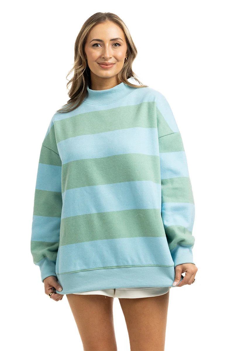 A woman stands facing forward and smiling, wearing the Katydid Aqua and Green Striped Mockneck Sweatshirt Women, paired with white shorts.