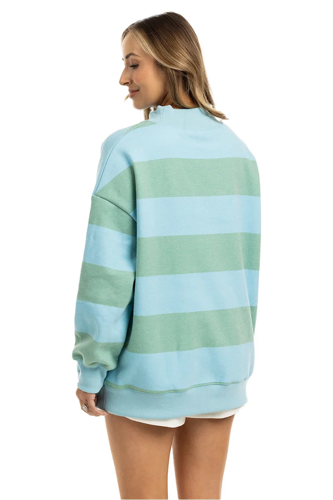 A woman with shoulder-length hair smiles slightly as she faces away, wearing the Katydid Aqua and Green Striped Mockneck Sweatshirt for women, styled with white shorts.