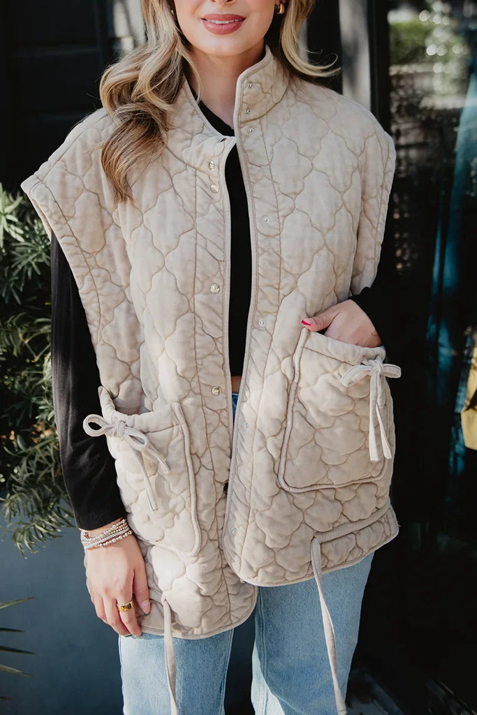 A woman stands outdoors in the Katydid Beige Oversized Garment Washed Vest, featuring large front pockets and a drawstring, styled over a black top and light blue jeans. Her loose hair and jewelry complete the look.