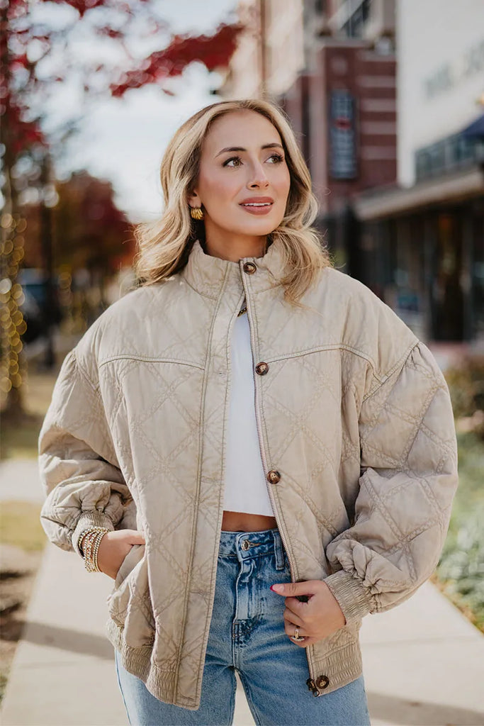 Outdoors on a sidewalk, a woman with blonde wavy hair and gold hoop earrings wears the Katydid Beige Luxe Oversized Jacket over a white crop top and blue jeans. She looks to the side, with fall trees and buildings in the background.