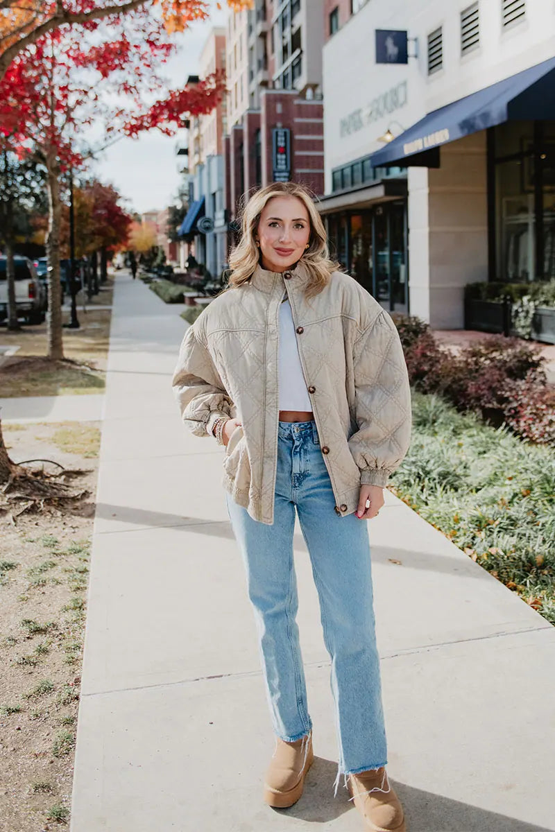 A woman stands on a city sidewalk wearing the Katydid Beige Luxe Oversized Jacket, paired with light blue jeans and tan shoes. Colorful autumn trees and shops are visible in the background.