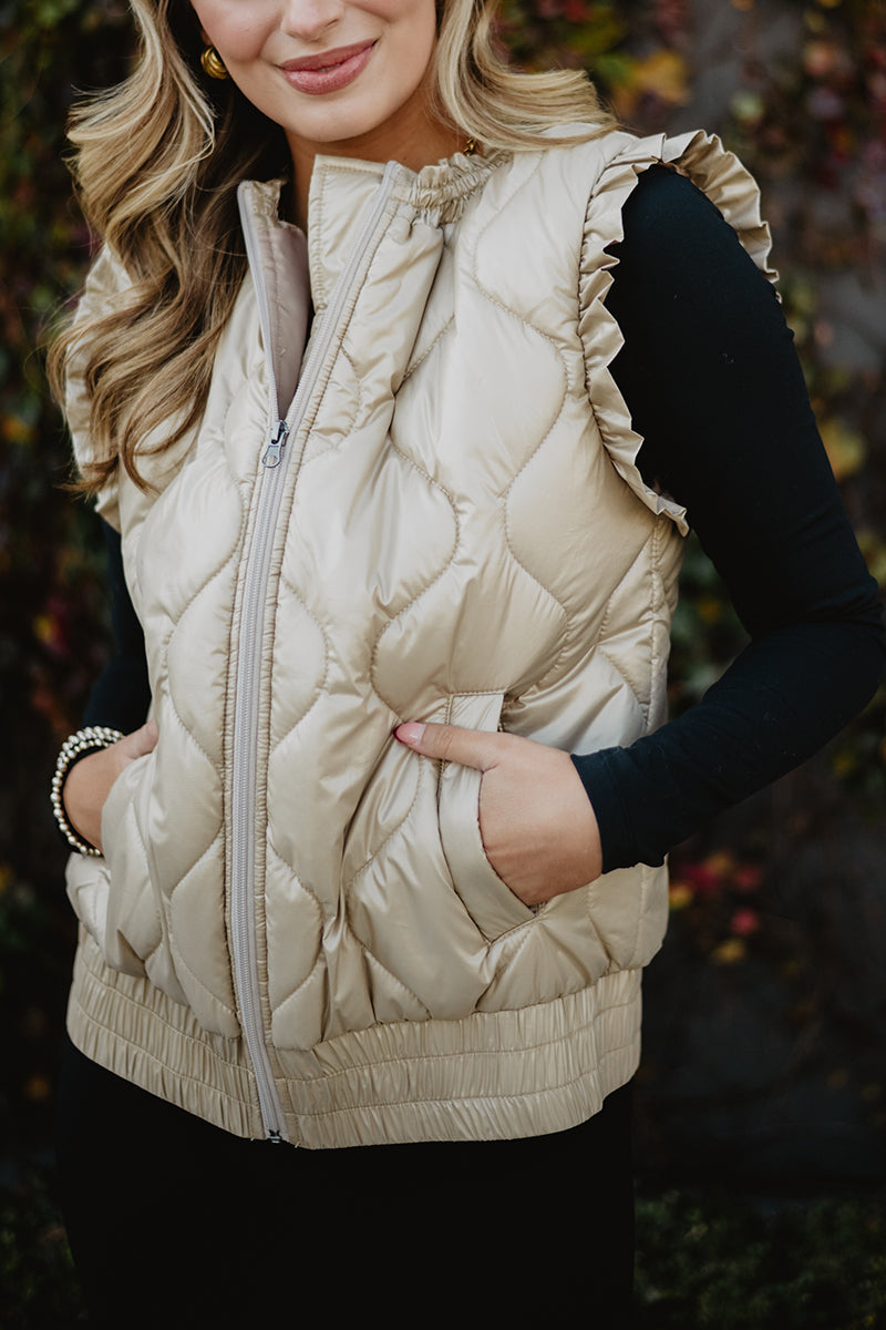A woman wearing the Katydid Soft Tan Metallic Quilted Vest over a long-sleeve black top smiles slightly with her hands in the vest pockets.