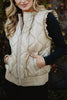 A woman wearing the Katydid Soft Tan Metallic Quilted Vest over a long-sleeve black top smiles slightly with her hands in the vest pockets.