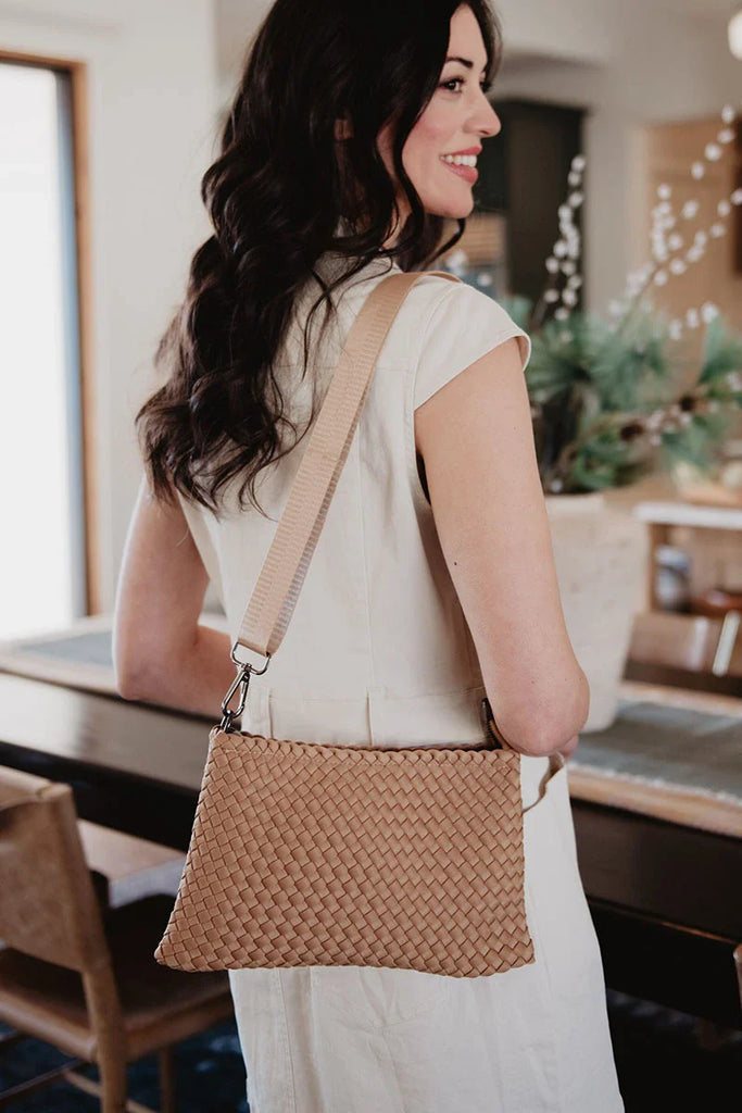 A woman with long dark hair, in a sleeveless light dress, smiles indoors while carrying the Katydid Camel Woven Everywhere Crossbody Sling Bag over her shoulder.