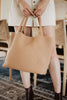 A woman stands indoors beside wooden chairs and a patterned rug, holding the Katydid Camel Woven Tote Bag by its handles, styled with a cream skirt and brown ankle boots.