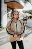 A woman smiles outdoors in front of a building, holding her Katydid Tan Sherpa Lace Trim Jacket closed. She styles the cozy zip-up with black leggings. Trees and patio umbrellas appear in the background.