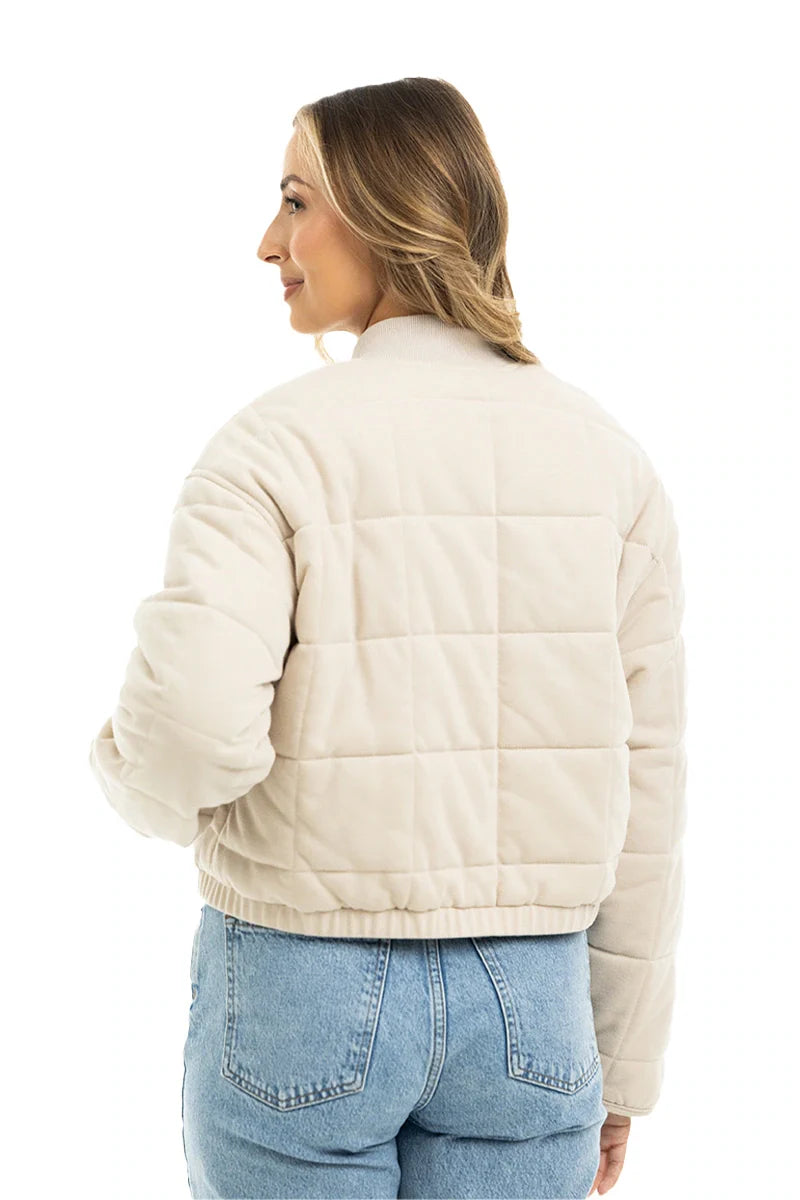 A woman with wavy blonde hair models the Katydid Beige Quilted Bomber Fall Jacket for women, paired with blue high-waisted jeans, as she faces away and looks over her shoulder against a plain white background.