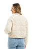 A woman with wavy blonde hair models the Katydid Beige Quilted Bomber Fall Jacket for women, paired with blue high-waisted jeans, as she faces away and looks over her shoulder against a plain white background.
