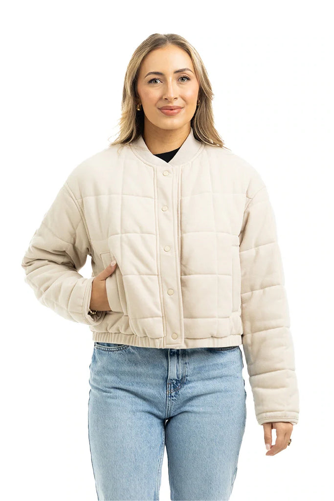 A woman wearing the Katydid Beige Quilted Bomber Fall Jacket with snap buttons and blue jeans stands against a white background, smiling.