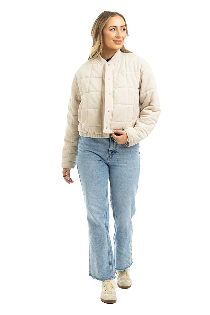 A woman wearing the Katydid Beige Quilted Bomber Fall Jacket, paired with light blue jeans and beige sneakers, stands against a white background and smiles gently while gazing to the side.