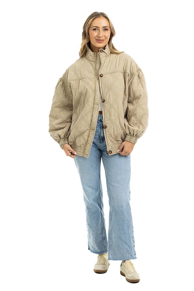 A woman smiles in the Katydid Beige Luxe Oversized Jacket, styled with light blue jeans and beige sneakers. Her blonde hair frames her face as she poses against a white background, highlighting the jacket’s comfort and style.