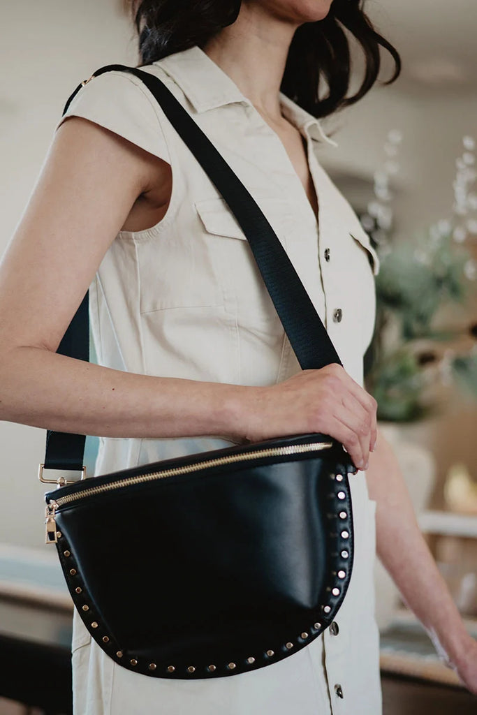 A woman in a sleeveless beige button-down dress carries the Katydid Black Gold-Studded Crossbody Bag, featuring metal studs and a gold zipper for an edgy touch. The image is cropped from her shoulders down.