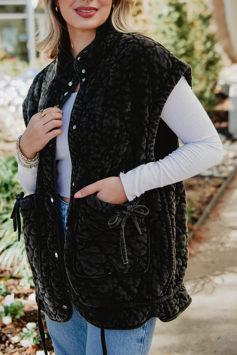 A woman in a white long-sleeve shirt, blue jeans, and a Katydid Charcoal Oversized Garment Washed Quilted Vest with front pockets stands on a path, holding the vest open with one hand and smiling slightly.