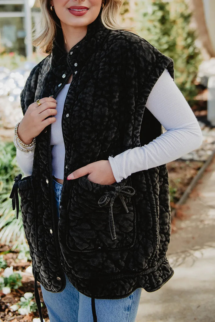A woman in a white long-sleeve shirt, blue jeans, and a Katydid Charcoal Oversized Garment Washed Quilted Vest with front pockets stands on a path, holding the vest open with one hand and smiling slightly.