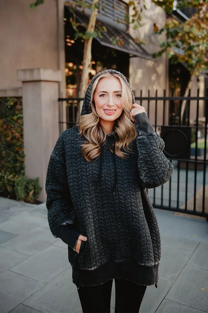 A woman with wavy blonde hair smiles outside on a paved sidewalk, wearing the Katydid Charcoal Heart Quilted Pullover Hoodie in front of a black metal fence and greenery.