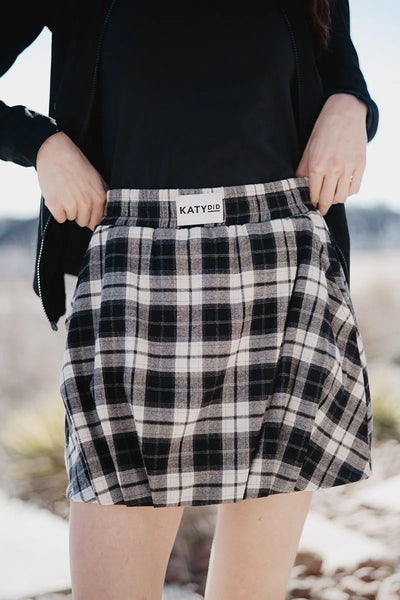 A person wears the Katydid Black Autumn Plaid Skirt with Shorts, paired with a black top and jacket outside against a blurred natural backdrop—making it an ideal autumn skirt choice.