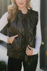 A woman models the Katydid Black Metallic Quilted Vest over a white long-sleeve top and black pants, accessorized with bracelets and rings, standing with one hand in her pocket near a white brick wall.