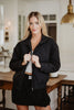 A woman with long blonde hair models the Katydid Black Sporty Diamond Quilted Jacket over a white top and black skirt, standing indoors near a table and brass light fixture.