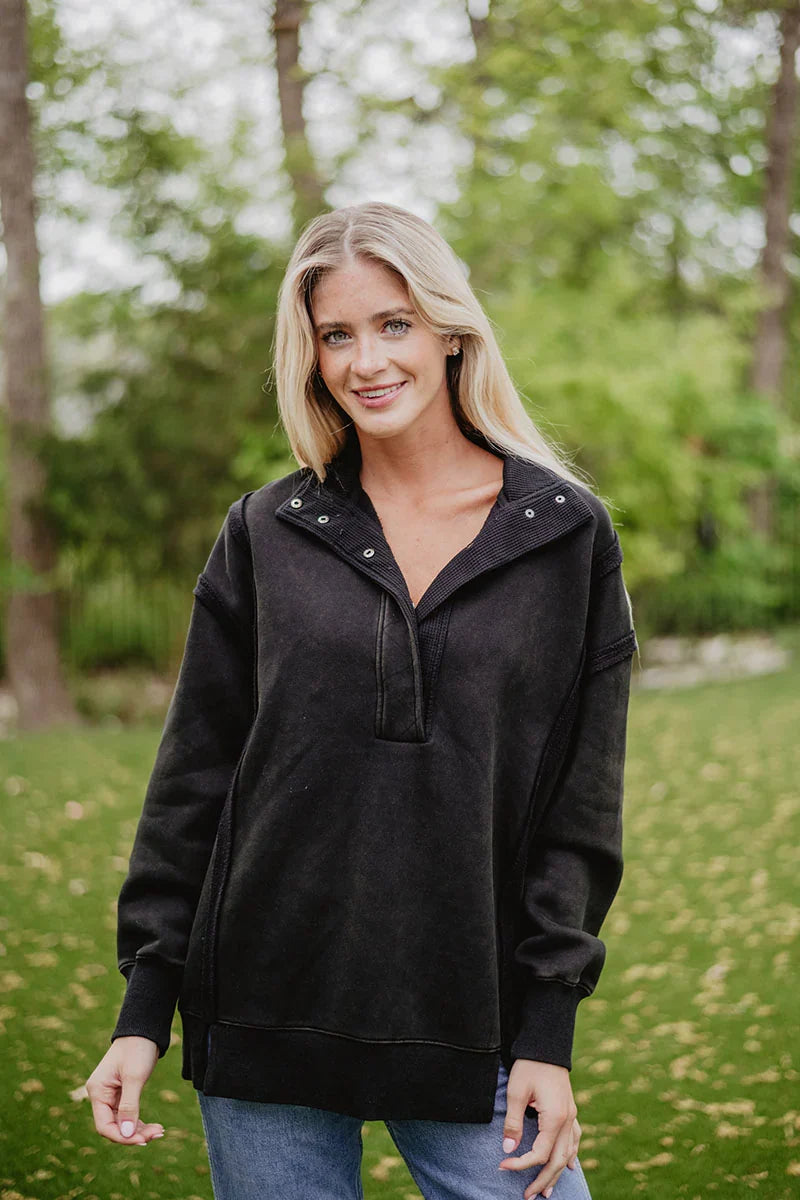 A woman with long blonde hair smiles at the camera outdoors, wearing the Katydid Black Vintage Wash Pullover Sweatshirt and blue jeans. Lush trees and greenery create a blurred backdrop.