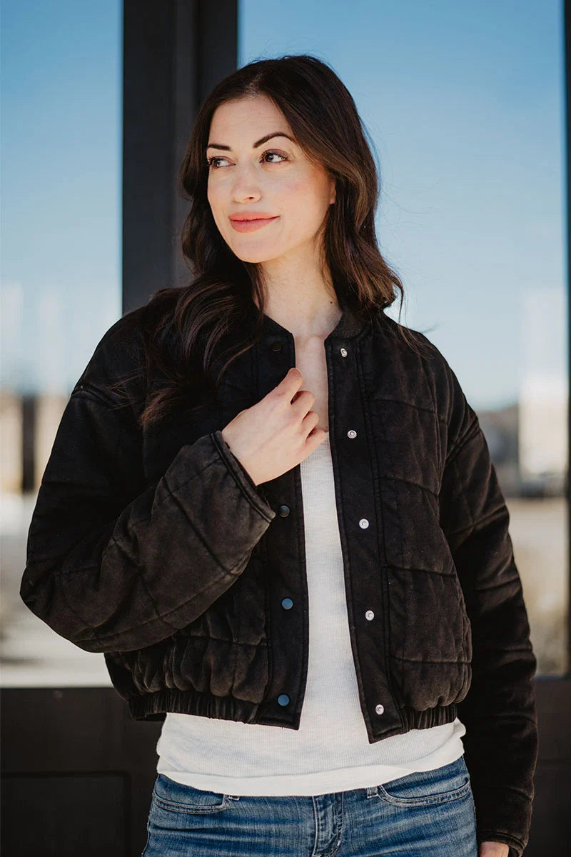 A woman with long brown hair wears the Katydid Black Cropped Quilted Bomber Jacket over a white shirt and blue jeans, standing outdoors by a glass door. This chic jacket brings warmth and elegance to her fall style as she smiles softly.
