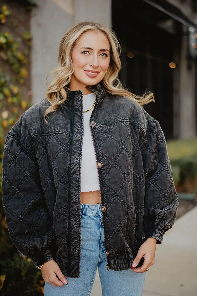 A woman with wavy blonde hair smiles outdoors in the Katydid Womens Charcoal Luxe Oversized Jacket, styled over a white crop top and blue jeans. One hand in her pocket, she strikes a vintage pose against lush greenery.