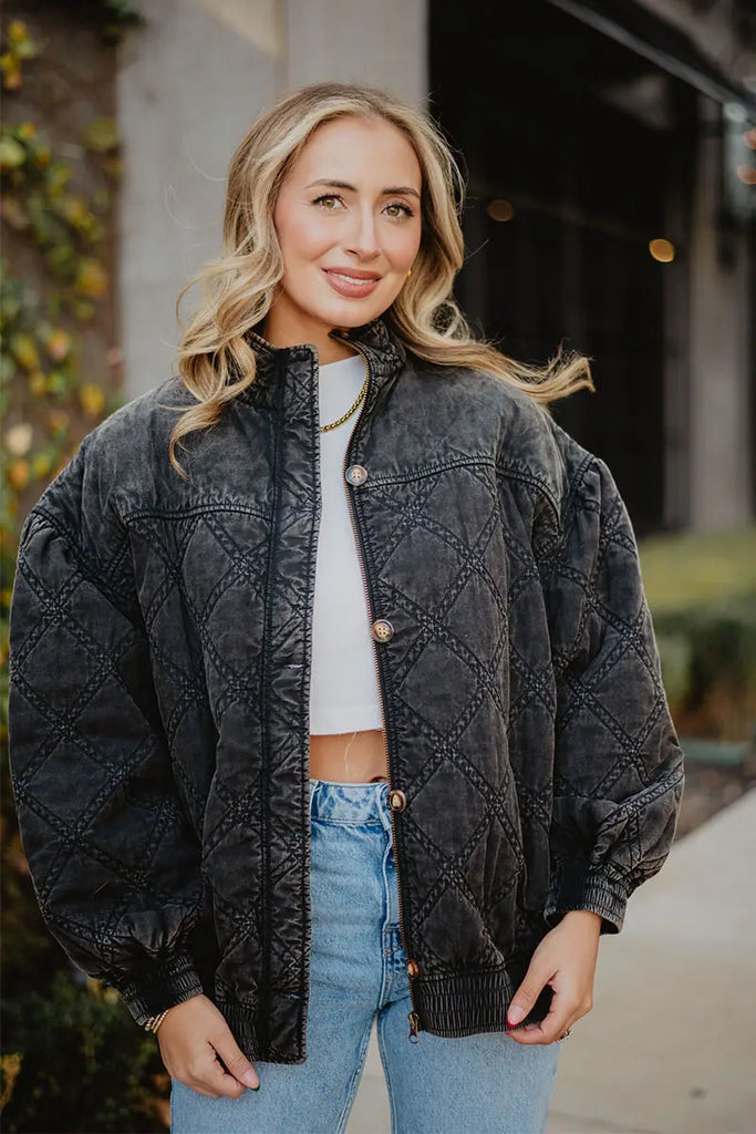 A woman with wavy blonde hair smiles outdoors in the Katydid Womens Charcoal Luxe Oversized Jacket, styled over a white crop top and blue jeans. One hand in her pocket, she strikes a vintage pose against lush greenery.