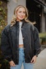 A woman with wavy blonde hair smiles outdoors in the Katydid Womens Charcoal Luxe Oversized Jacket, styled over a white crop top and blue jeans. One hand in her pocket, she strikes a vintage pose against lush greenery.
