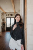 A woman with long brown hair smiles with her eyes closed, leaning against a stone wall in a bright hallway as she wears the Katydid Black Sporty Diamond Quilted Jacket paired with white shorts.