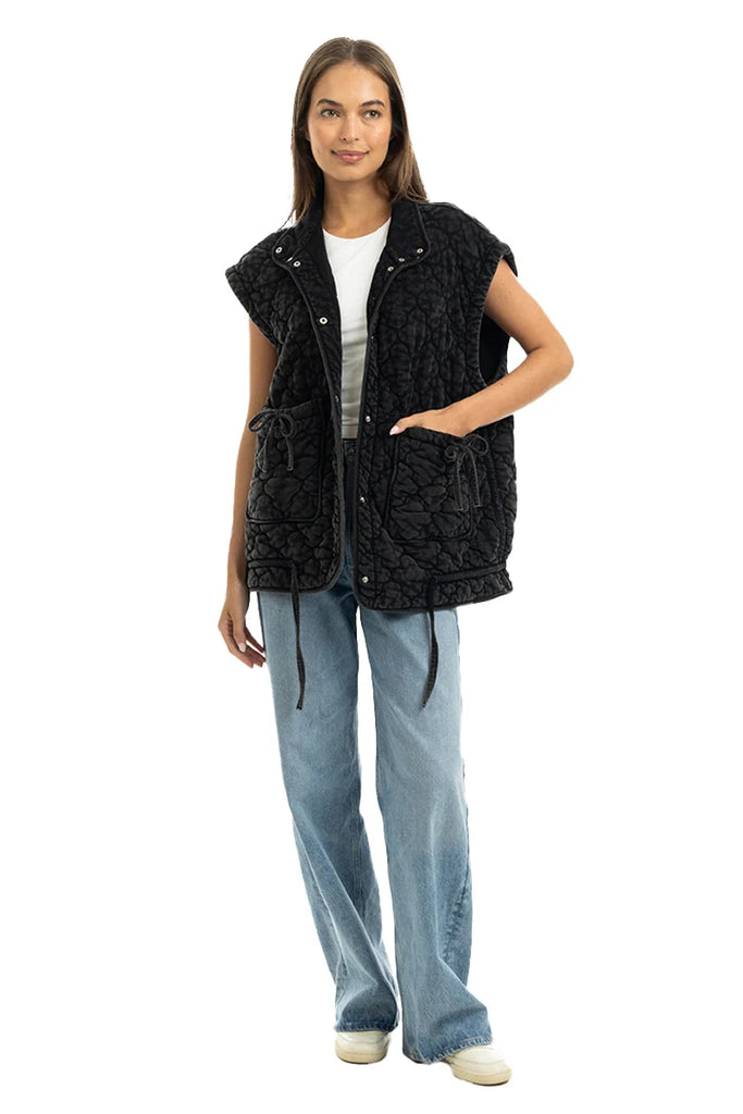A woman smiles in casual attire, wearing the Katydid Charcoal Oversized Garment Washed Quilted Vest over a white shirt, paired with light blue wide-leg jeans and white shoes against a plain white background.