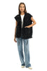 A woman smiles in casual attire, wearing the Katydid Charcoal Oversized Garment Washed Quilted Vest over a white shirt, paired with light blue wide-leg jeans and white shoes against a plain white background.