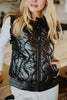 A woman with loose blonde hair stands indoors, partially smiling with her hands in the pockets of a Katydid Black Metallic Quilted Vest worn over a white long-sleeve shirt—a stylish women’s fall layer for cooler days.