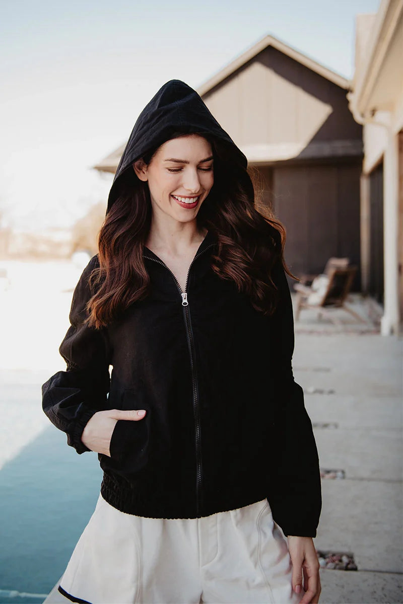 A woman with long brown hair stands by a pool, smiling and looking down with one hand in her pocket, wearing white shorts and the Katydid Black Lightweight Hooded Cute Jacket.