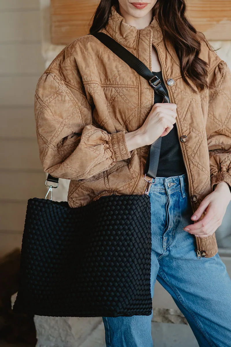 A woman in a brown quilted jacket and blue jeans stands holding the Katydid Black Woven Design Tote, which features a large size and wide strap.