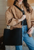 A woman in a brown quilted jacket and blue jeans stands holding the Katydid Black Woven Design Tote, which features a large size and wide strap.