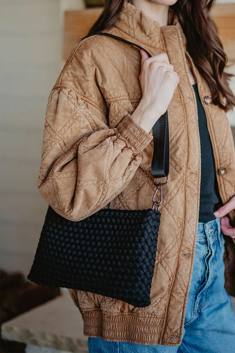 A person in a tan quilted jacket and blue jeans holds the Katydid Black Woven Everywhere Crossbody Women with a wide strap. The persons face is not visible.
