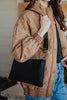 A person in a tan quilted jacket and blue jeans holds the Katydid Black Woven Everywhere Crossbody Women with a wide strap. The persons face is not visible.