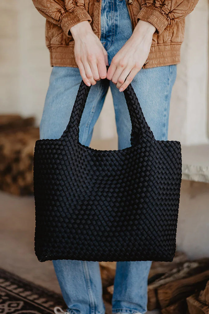 A person in a brown jacket and blue jeans holds the Katydid Black Woven Design Tote, a chic womens handbag, with both hands in front.