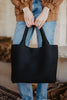 A person in a brown jacket and blue jeans holds the Katydid Black Woven Design Tote, a chic womens handbag, with both hands in front.