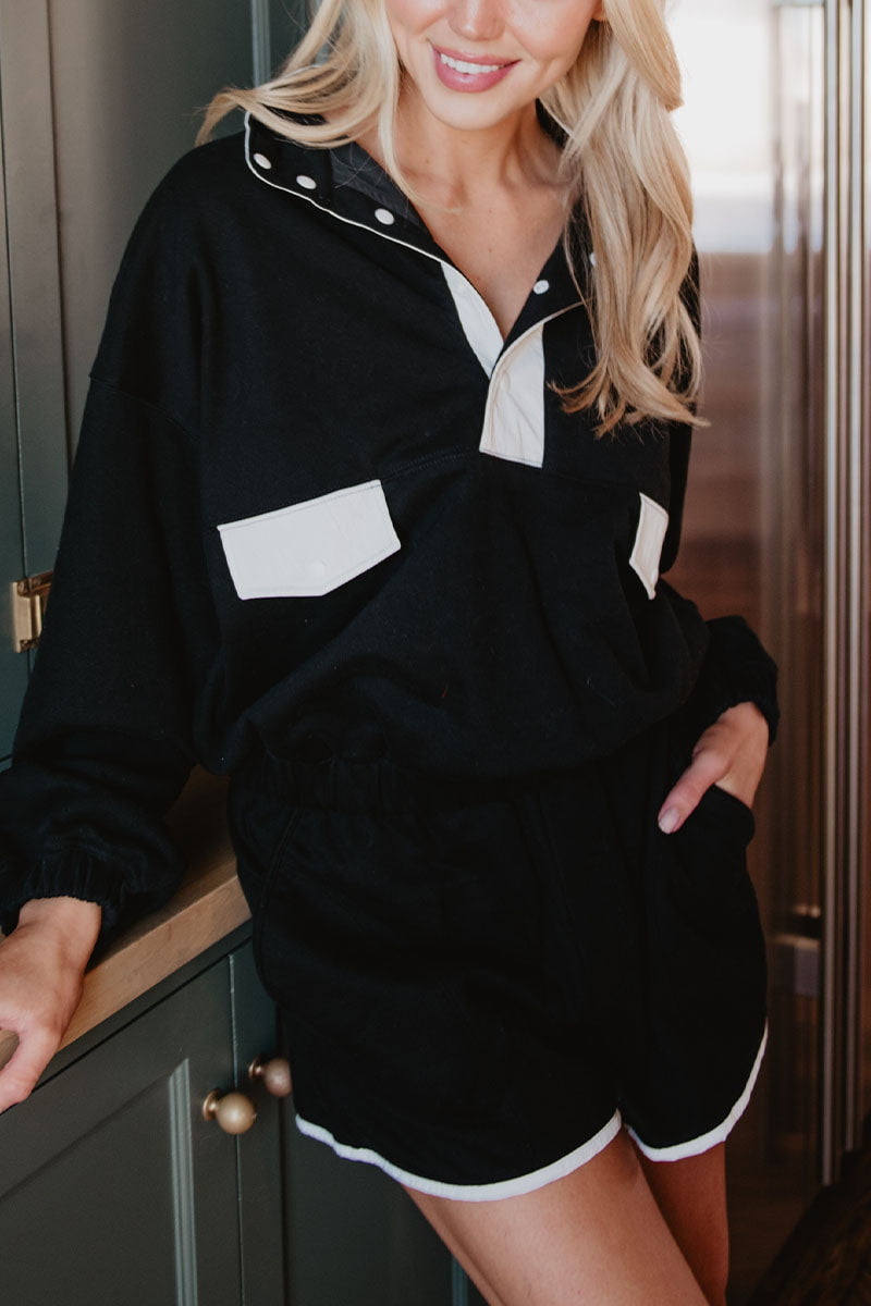 A woman stands indoors, smiling confidently with one hand in her pocket, wearing Katydids Black  White Quarter Snap Athletic Set for women—a sporty pullover and matching black shorts trimmed with white.