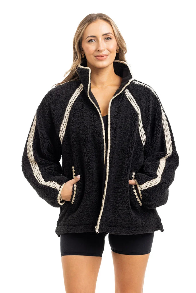 A woman wears the Katydid Black Sherpa Lace Trim Jacket, featuring ivory lace accents and front pockets, styled over a black top and shorts. She stands facing forward against a white background.