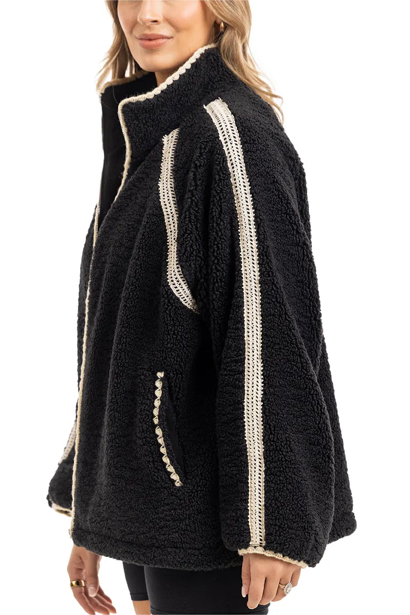A woman stands in profile, smiling slightly, wearing the Katydid Black Sherpa Lace Trim Jacket featuring cozy fabric and cream crochet detailing on the shoulders, arms, and pockets.