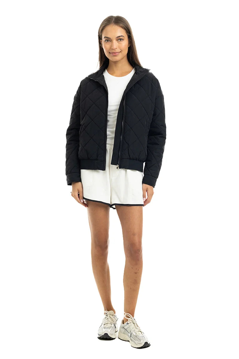 A woman with straight brown hair and a neutral expression models the Katydid Black Sporty Diamond Quilted Jacket over a white dress and sneakers, standing against a plain white background.