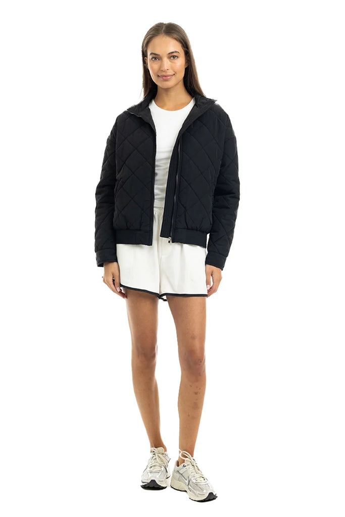 A woman with straight brown hair and a neutral expression models the Katydid Black Sporty Diamond Quilted Jacket over a white dress and sneakers, standing against a plain white background.