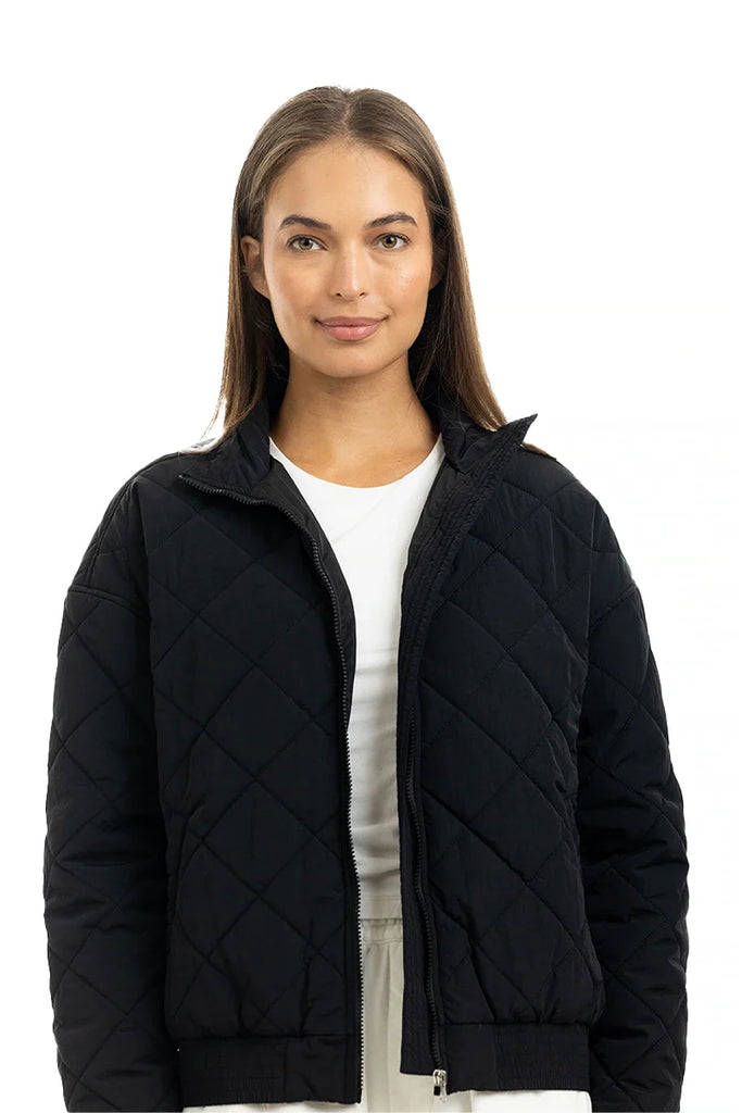 A woman with straight brown hair wears a white shirt and the Katydid Black Sporty Diamond Quilted Jacket, standing against a plain white background and smiling gently at the camera—ideal as a stylish fall jacket.