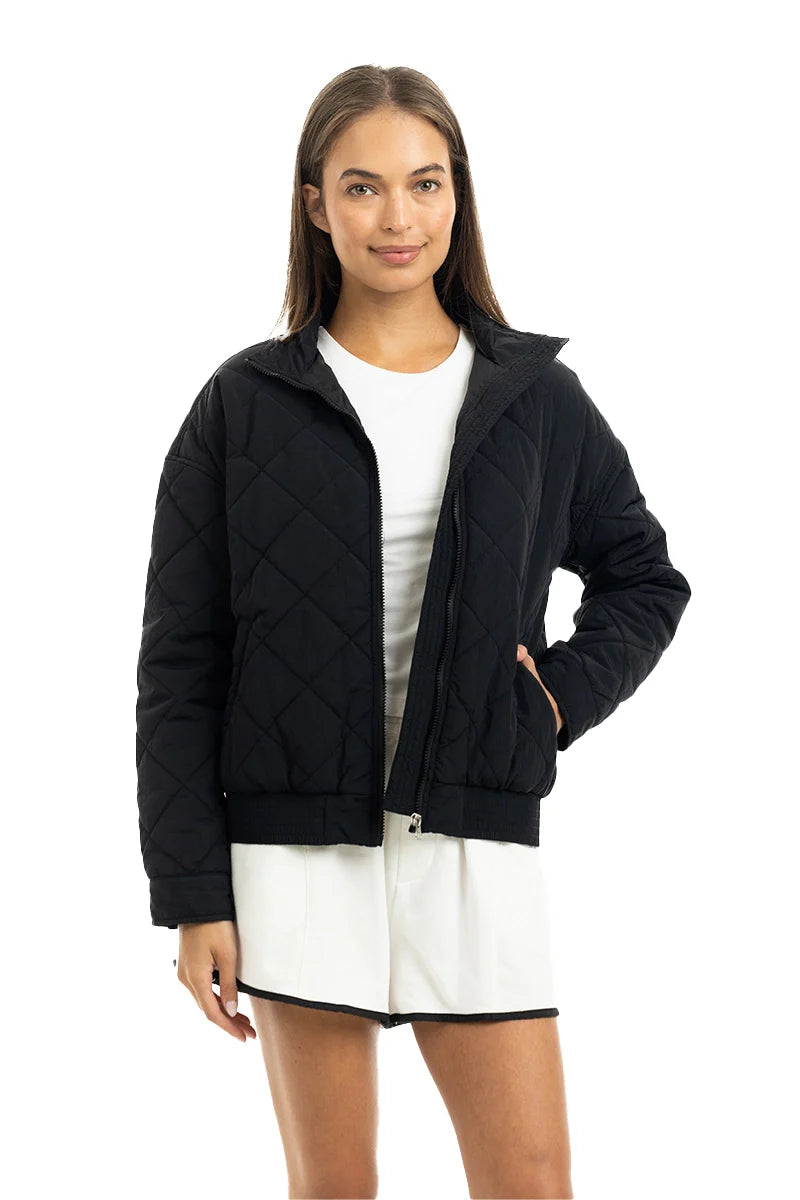 A person wearing Katydids Black Sporty Diamond Quilted Jacket, styled over a white dress, stands against a plain white background and smiles slightly.