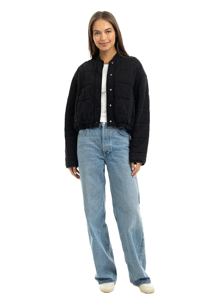 A woman faces forward against a white background, wearing the Katydid Black Cropped Quilted Bomber Jacket over a white shirt, paired with light blue jeans and white sneakers.