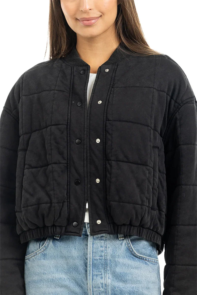A person models the Katydid Black Cropped Quilted Bomber Jacket with snap buttons over a white top and light blue jeans, shown from shoulders to waist against a white background that highlights this classic fall style.