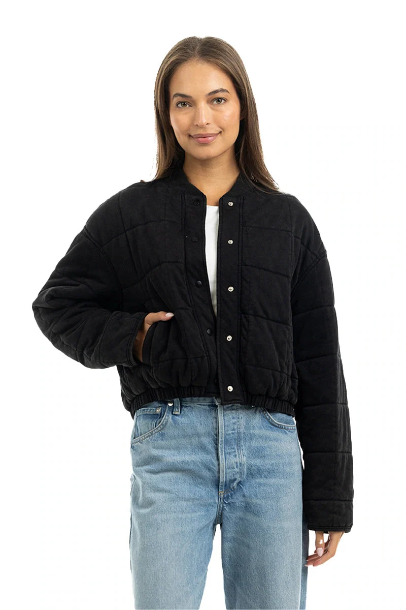 A woman with long brown hair wears the Katydid Black Cropped Quilted Bomber Jacket over a white shirt and light blue jeans, one hand in her pocket, smiling at the camera against a white backdrop—an ideal fall look.