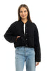A woman with long brown hair wears the Katydid Black Cropped Quilted Bomber Jacket over a white shirt and light blue jeans, one hand in her pocket, smiling at the camera against a white backdrop—an ideal fall look.